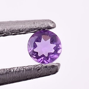 Puede incluir: Una piedra preciosa de color púrpura en forma de estrella sostenida por unas pinzas. La piedra preciosa es redonda y tiene un corte facetado.