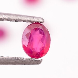 Può includere: Primo piano di una gemma ovale di colore rosso brillante tenuta in una pinza. La gemma è circondata da uno sfondo bianco.