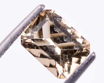 May include: A light brown gemstone with a rectangular shape, held by tweezers against a white background.