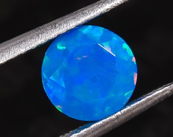 May include: A round blue opal gemstone with iridescent flashes of color, held in tweezers against a black background.