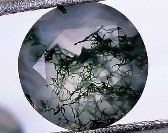 May include: A round, green and white agate gemstone with a mossy pattern. The gemstone is held by a pair of tweezers.