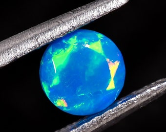 May include: A blue opal gemstone with green and yellow flashes of color is held by silver tweezers against a black background.