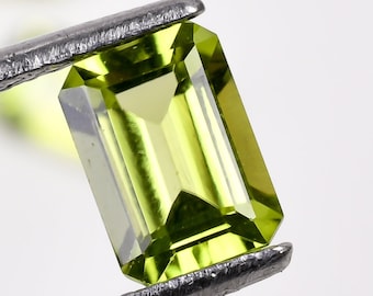 May include: A green emerald cut gemstone held in tweezers. The gemstone is a vibrant green color and has a smooth, polished surface.