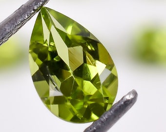 May include: A pear-shaped green peridot gemstone held by tweezers. The gemstone is faceted and has a bright, vibrant green color.