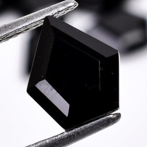 May include: A black gemstone with a faceted, kite-shaped cut, held by tweezers. The gemstone is a dark, opaque black.