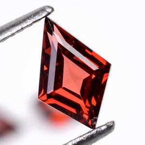 Puede incluir: Una piedra preciosa roja con forma de cometa está sujeta por unas pinzas. La piedra preciosa está facetada y tiene un alto brillo.