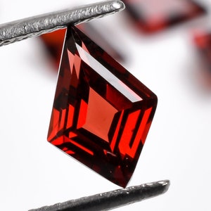Könnte beinhalten: Ein roter Edelstein mit einem facettierten, drachenförmigen Schliff wird von einer Pinzette gehalten. Der Edelstein ist von anderen roten Edelsteinen umgeben.