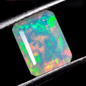 Puede incluir: Una piedra preciosa de ópalo blanco con destellos verdes y naranjas, sostenida por unas pinzas sobre un fondo negro.