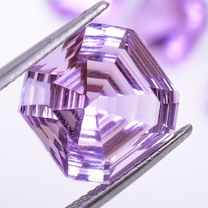 May include: A purple gemstone with a faceted cut, held by tweezers. The gemstone is a hexagonal shape with a clear, light purple color.