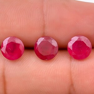 May include: Three round, faceted ruby gemstones in a deep red color. The gemstones are approximately 5mm in diameter and are displayed on a hand.