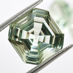 May include: A light green gemstone with a faceted cut, held by tweezers. The gemstone is a square shape with rounded edges.