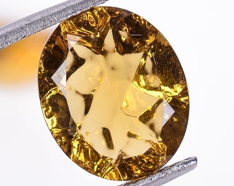 May include: A golden yellow oval-shaped gemstone with a faceted cut. The gemstone is held in place by a pair of tweezers.