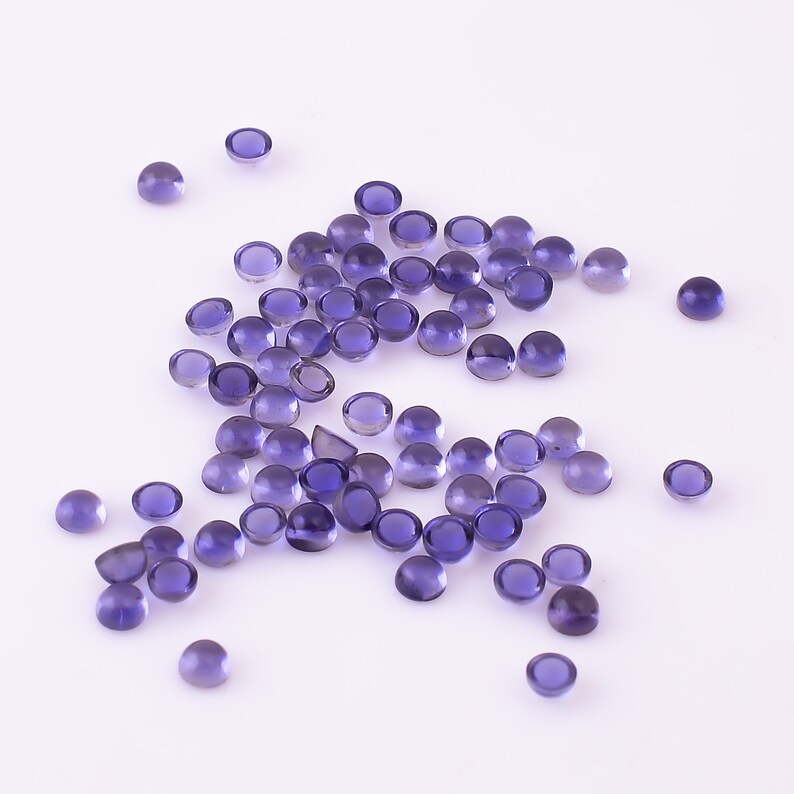 May include: A collection of round, purple gemstones, also known as amethyst, scattered on a white surface. The gemstones are smooth and polished, with a slight sheen.