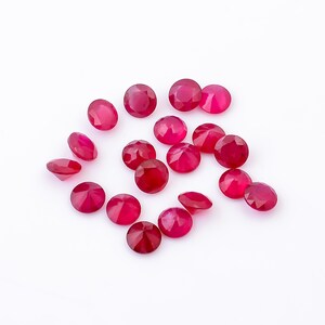 May include: A collection of 15 round, faceted ruby gemstones. The gemstones are a deep red color and are scattered on a white surface.