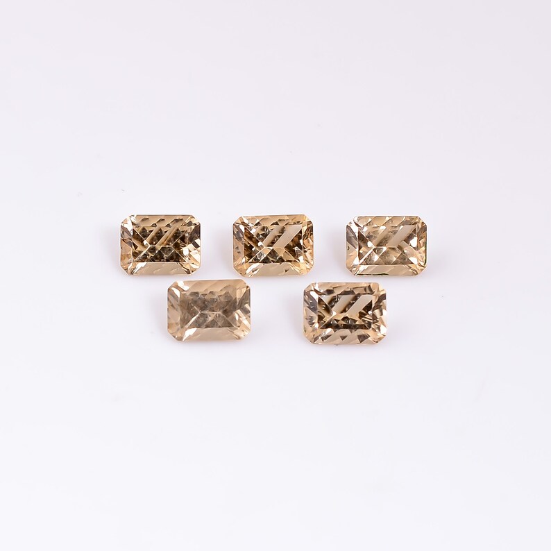 May include: Five light brown, faceted, rectangular gemstones on a white background.