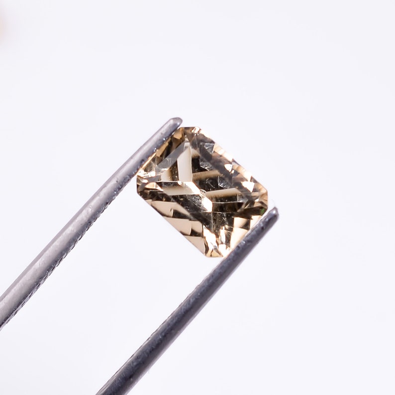 May include: A light brown gemstone with a rectangular shape, held by tweezers against a white background.
