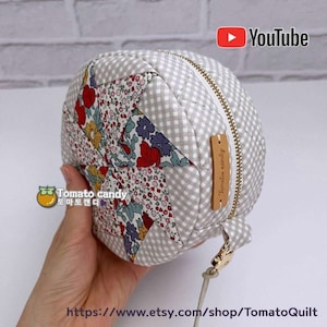May include: A round, gray and white fabric bag with a zipper closure and a small brown leather tag that says "Tomato candy". The bag is decorated with a patchwork design of floral fabric.