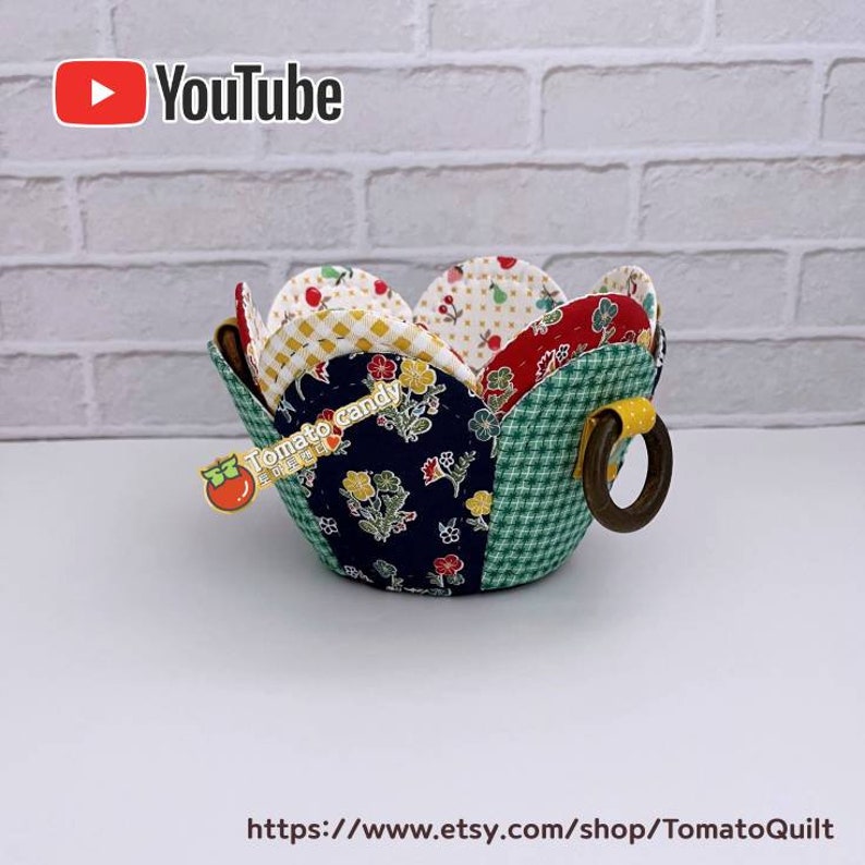 Peut inclure: Un bol en forme de fleur en tissu patchwork avec une poign&eacute;e en anneau en bois. Le bol est fait de diff&eacute;rents tissus color&eacute;s avec des motifs floraux et g&eacute;om&eacute;triques. Le bol est pos&eacute; sur une surface blanche.