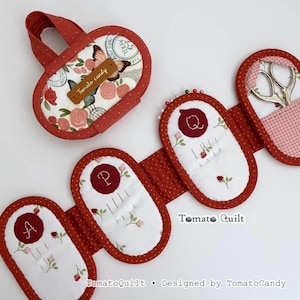 Puede incluir: Un estuche de tela para agujas con borde de lunares rojos y estampado floral que incluye una mariposa y el texto Tomato candy. El estuche se despliega para mostrar tres secciones ovaladas con las letras A, P y Q bordadas y un bolsillo con tijeras.