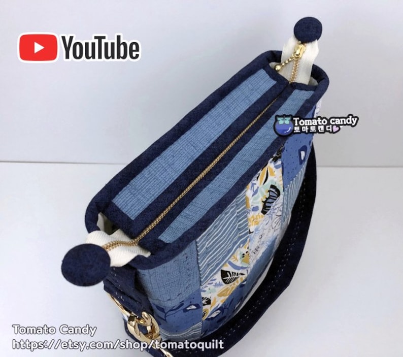 Pode incluir: Uma bolsa tote de tecido patchwork azul e branco com um z&iacute;per dourado e uma al&ccedil;a azul marinho. A bolsa tem um pequeno remendo redondo azul marinho com o texto "Tomato candy" em branco e um cora&ccedil;&atilde;o rosa.