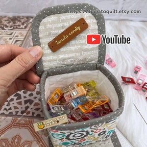 May include: A gray fabric box with a white polka dot pattern and floral print on the bottom. The box is open and filled with colorful plastic clips. The box has a brown leather tag that says "Tomato candy" and a white tag that says "Tomato candy 토마토캔디".