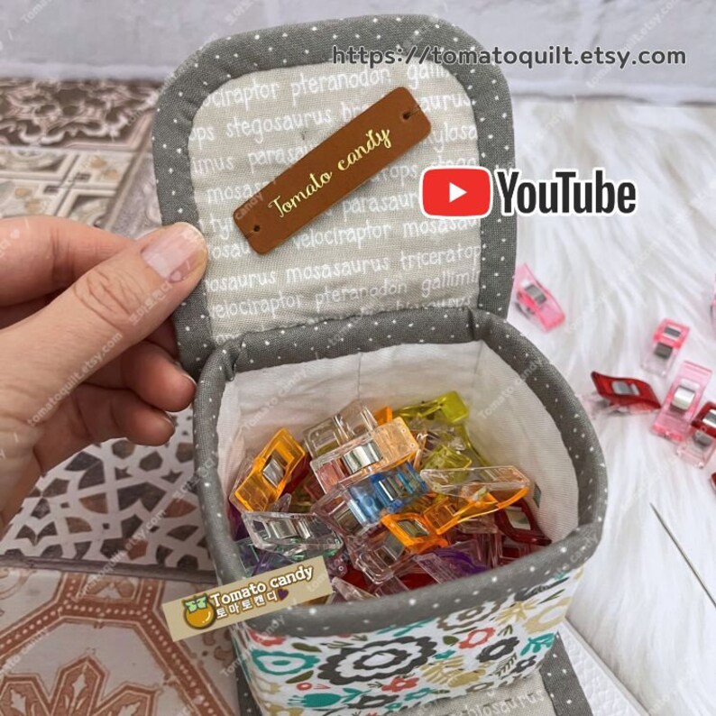 May include: A gray fabric box with a white polka dot pattern and floral print on the bottom. The box is open and filled with colorful plastic clips. The box has a brown leather tag that says "Tomato candy" and a white tag that says "Tomato candy 토마토캔디".