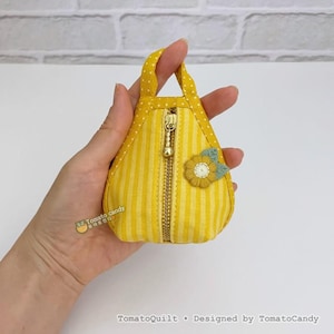 May include: A small, yellow fabric bag with a vertical striped pattern and a gold zip. The bag has a small handle and a decorative flower appliqué. The text "TomatoQuilt • Designed by TomatoCandy" is at the bottom.