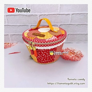 Peut inclure: Un contenant de rangement rond en tissu matelassé avec un motif patchwork dans des tons de rouge, jaune et blanc. Il comprend une poignée jaune, un couvercle zippé avec deux pompons et une étiquette brune avec le texte Tomato candy.