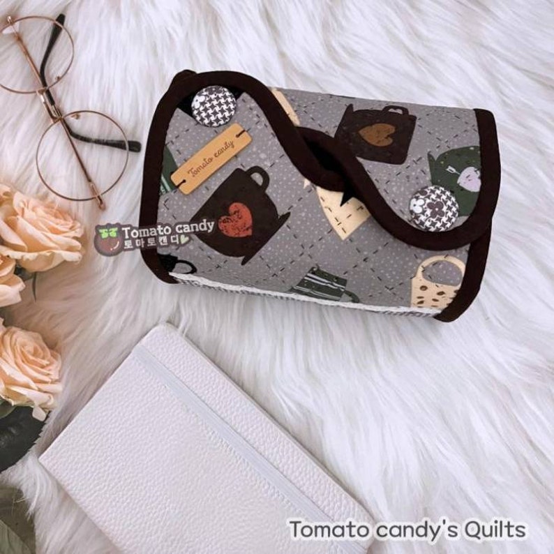May include: A gray quilted fabric pouch with a brown trim and a brown label that says "Tomato candy". The pouch is decorated with a pattern of teacups and hearts. The pouch is sitting on a white fluffy surface.