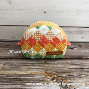 May include: A semi-circular quilted pouch featuring a diamond-patterned patchwork design in shades of orange, yellow, green, and white. A brown leather-like tag on the front is embossed with the text Tomato candy. The pouch has small side tabs.