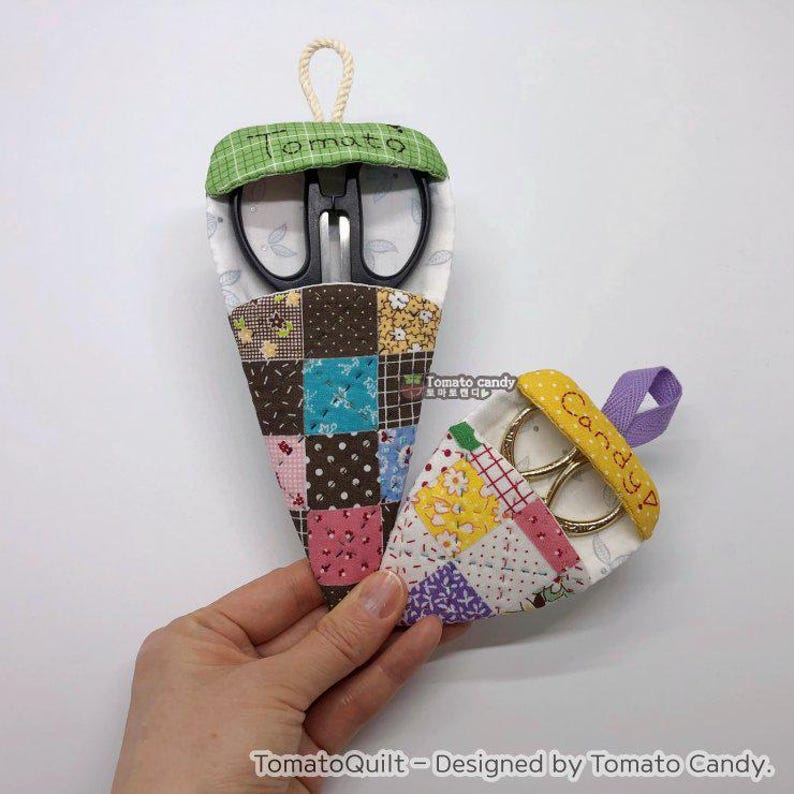 May include: Two fabric pouches with embroidered names "Tomato" and "Candy!"  Each pouch is made of patchwork fabric and holds a pair of scissors. The pouches are hanging from a white background.