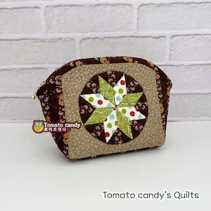 Puede incluir: Un estuche de tela acolchada con estampado floral marrón oscuro y un parche circular central con un diseño de estrella multicolor en verde, blanco y rojo. El estuche tiene un tirador de cremallera dorado y el logo Tomato candy.