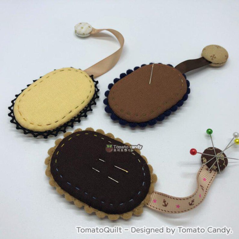May include: Three fabric pincushions with different colored fabric and trim. Each pincushion has a ribbon attached to it. The pincushions are shaped like an oval and have a decorative trim around the edge. The pincushions are in the colors of brown, yellow, and blue.