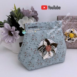 May include: A light blue fabric pouch with a floral print and a brown and white fabric flower with a pink center. The pouch has a zipper closure and the text "Tomato candy" is printed on the front.
