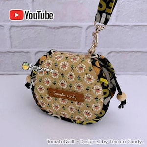 May include: A quilted pouch featuring a tan fabric with a repeating white and pink floral pattern. A brown rectangular label with the text Tomato candy is centred on the front. The pouch has black trim, wooden beads, and a patterned strap.