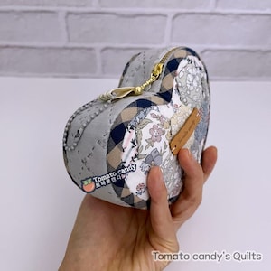 Peut inclure: Une pochette en tissu de forme cœur avec une fermeture à glissière et une petite étiquette en cuir qui indique "Tomato candy". La pochette est faite de différents tissus, dont des imprimés floraux, du plaid et des couleurs unies. La pochette est tenue dans une main.