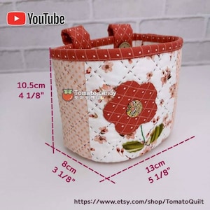 May include: A quilted fabric storage basket with a floral print and a red flower applique. The basket has two handles and measures 4 1/8 inches tall, 3 1/8 inches wide, and 5 1/8 inches long.