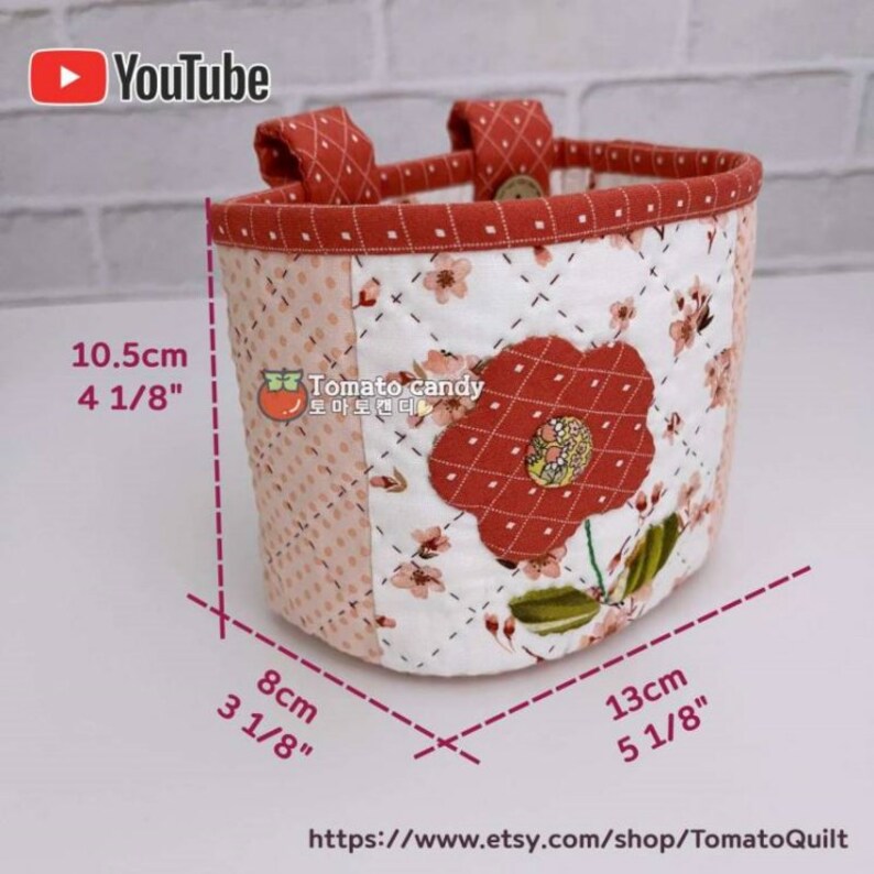 May include: A quilted fabric storage basket with a floral print and a red flower applique. The basket has two handles and measures 4 1/8 inches tall, 3 1/8 inches wide, and 5 1/8 inches long.