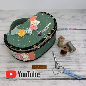 May include: A green fabric half-circle box with a zipper closure and a handle. The box is decorated with colorful patchwork fabric and has a small label that says "Tomato candy". The box is sitting on a wooden surface with a pair of scissors, spools of thread, and a blue tag with the word "SAJOU" on it.
