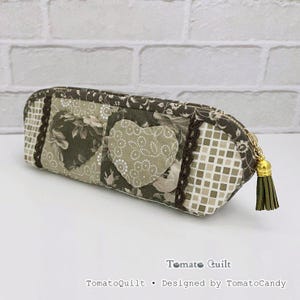 Può includere: Un astuccio rettangolare in tessuto con un design patchwork di motivi floreali verde oliva e beige, applicazioni a forma di cuore e una stampa a griglia. Include un bordo in pizzo marrone scuro, una cerniera dorata e una nappa.