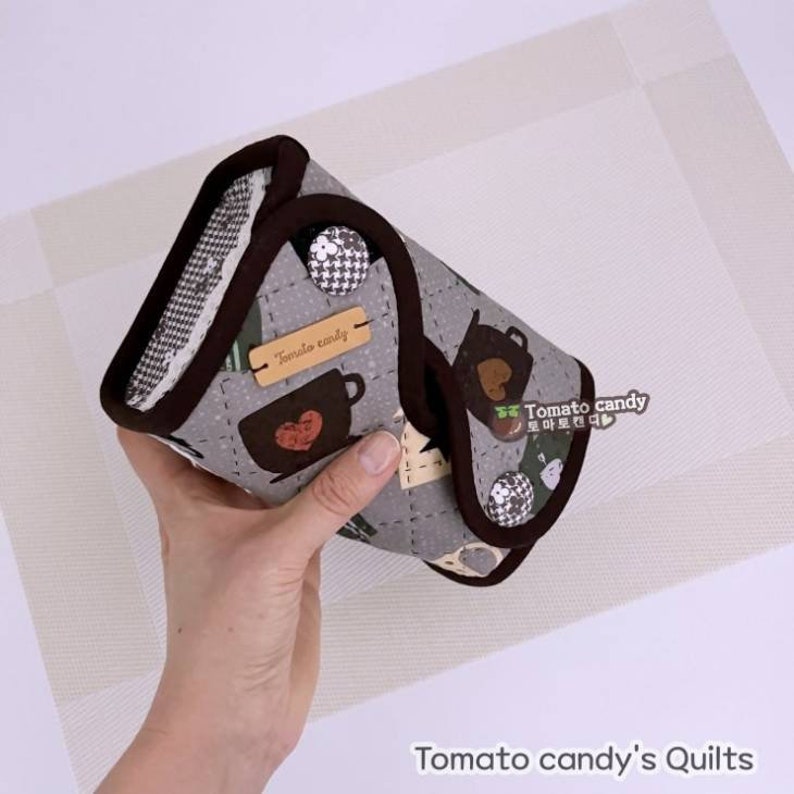 May include: A gray and brown quilted fabric pouch with a brown trim and a brown button. The pouch is decorated with a pattern of teacups and hearts. The pouch is labeled "Tomato candy".