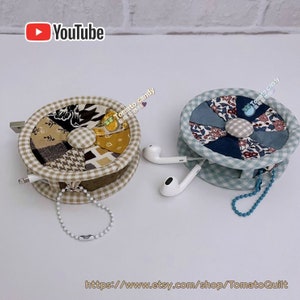 Pode incluir: Dois organizadores de cabos feitos à mão de tecido, um em bege e branco com um padrão xadrez e o outro em azul e branco com um padrão floral. Ambos os organizadores têm um pequeno botão no centro e uma corrente presa ao lado. Os organizadores estão cheios de fones de ouvido brancos.