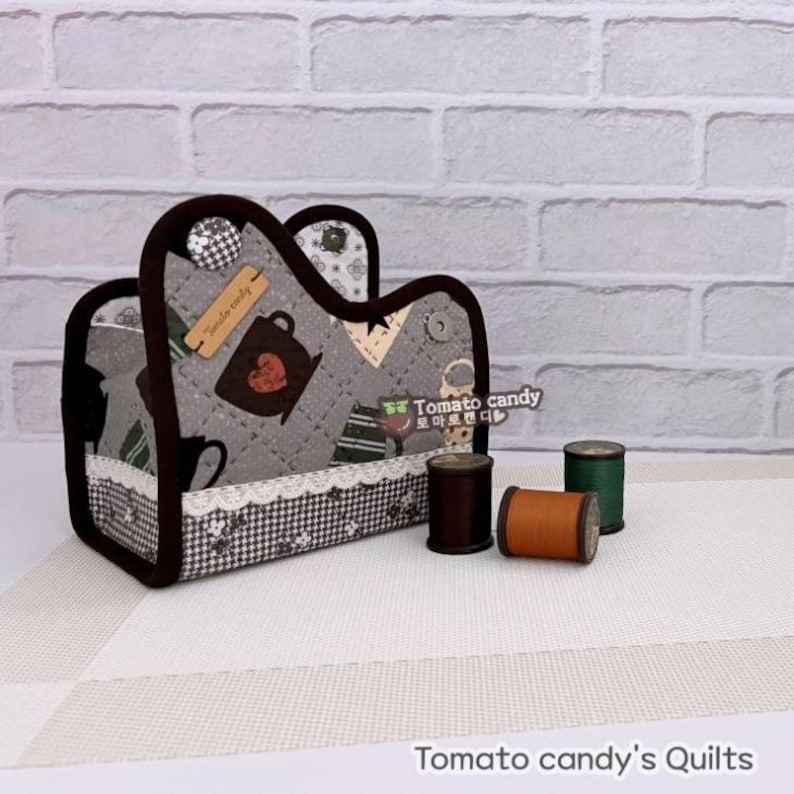 May include: A gray and brown quilted fabric storage box with a lace trim and a brown button. The box has a cutout in the shape of a teacup and the text "Tea Time Today" on a tag. The box is sitting on a white surface with three spools of thread.