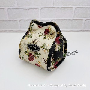 May include: A floral print fabric pouch with a black trim. The pouch features a cream-colored background with a pattern of red, white, and green flowers. The text "Tomato Candy" is on a small black label.