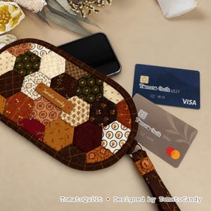 May include: A quilted fabric pouch featuring a hexagonal patchwork pattern in shades of brown, cream, and burgundy. A brown leather tag labeled Tomato candy is attached. The pouch includes a wrist strap and contains a smartphone and two credit cards.