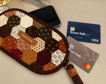 May include: A quilted fabric pouch featuring a hexagonal patchwork pattern in shades of brown, cream, and burgundy. A brown leather tag labeled Tomato candy is attached. The pouch includes a wrist strap and contains a smartphone and two credit cards.