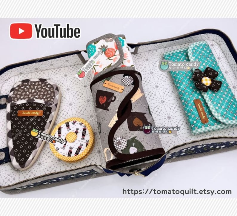 May include: An open gray and blue fabric case with various fabric pouches and accessories. The pouches are decorated with different patterns and designs, including a donut, a cup of coffee, and a flower. The case has a zipper closure and a label that reads "Tomato Candy".