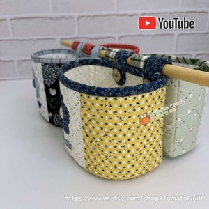May include: Three fabric hanging baskets with a wooden dowel rod. The baskets are made of patchwork fabric in various patterns, including yellow with black polka dots, black and white, and green with white polka dots. The baskets are hanging from the dowel rod with blue and white fabric loops. The text "Tomato candy" is visible on the yellow basket.