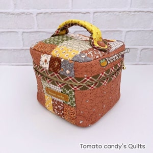 May include: A square fabric storage box with a patchwork lid featuring floral and geometric patterns in shades of brown, green, and yellow. It has a yellow handle, a zippered closure, and a brown leather tag labeled Tomato candy.