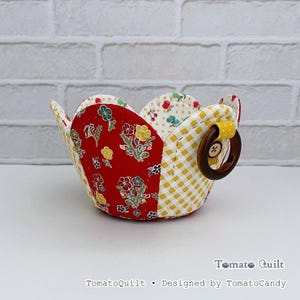 May include: A fabric bowl featuring a scalloped edge with alternating panels of red floral print, yellow and white gingham, and a cream cherry print. A dark brown wooden ring is attached to one side with a yellow polka dot fabric tab. Text reads TomatoQuilt.